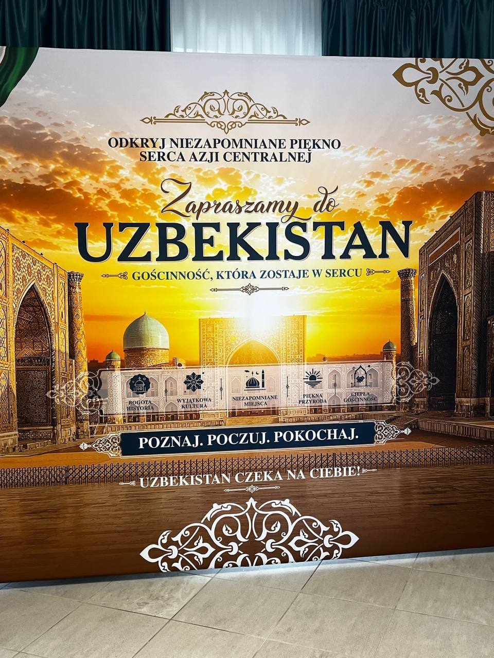 Promocyjna ścianka o Uzbekistanie z hasłem zachęcającym do odkrywania kraju oraz grafiką przedstawiającą tradycyjną uzbecką architekturę.